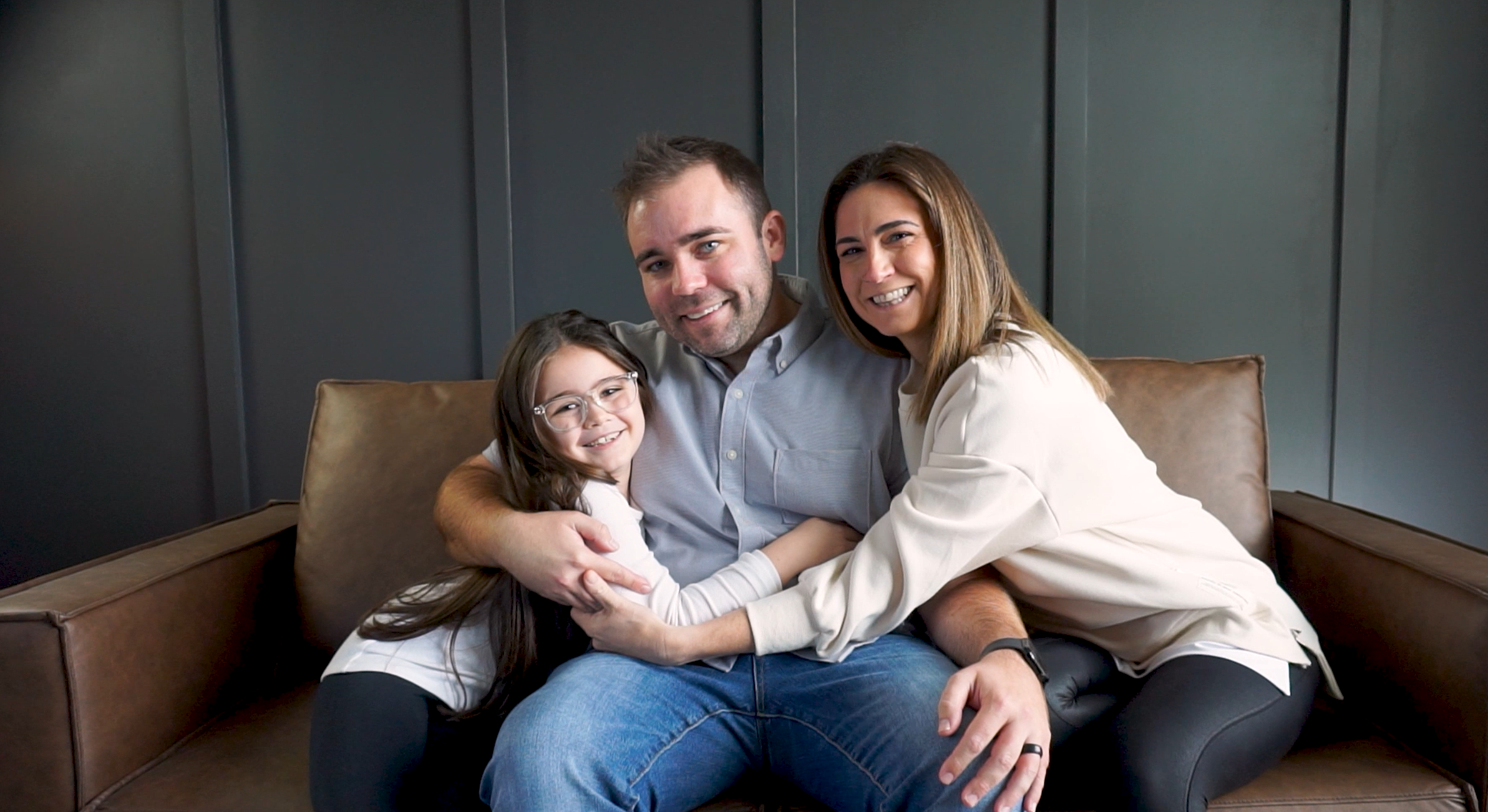 young family of three sitting on brown couch and hugging