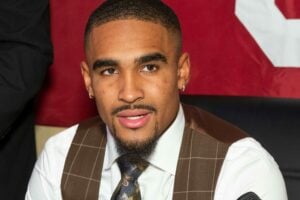 Jalen Hurts seated at a press event, wearing a white shirt, brown plaid vest, and patterned tie, with a microphone positioned in front of him.
